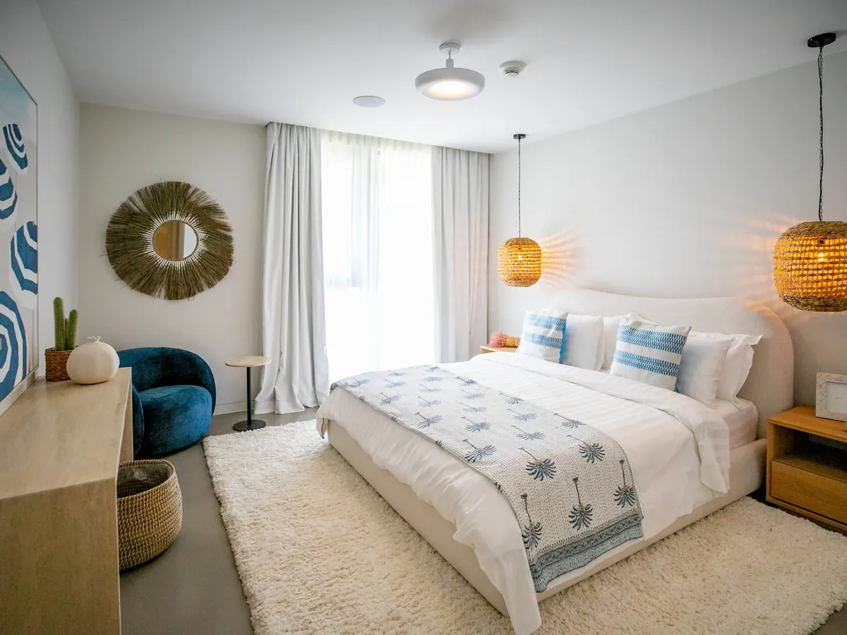 Bright Dubai apartment bedroom with white bed linen, sheer curtains, woven pendant light and a round gold mirror