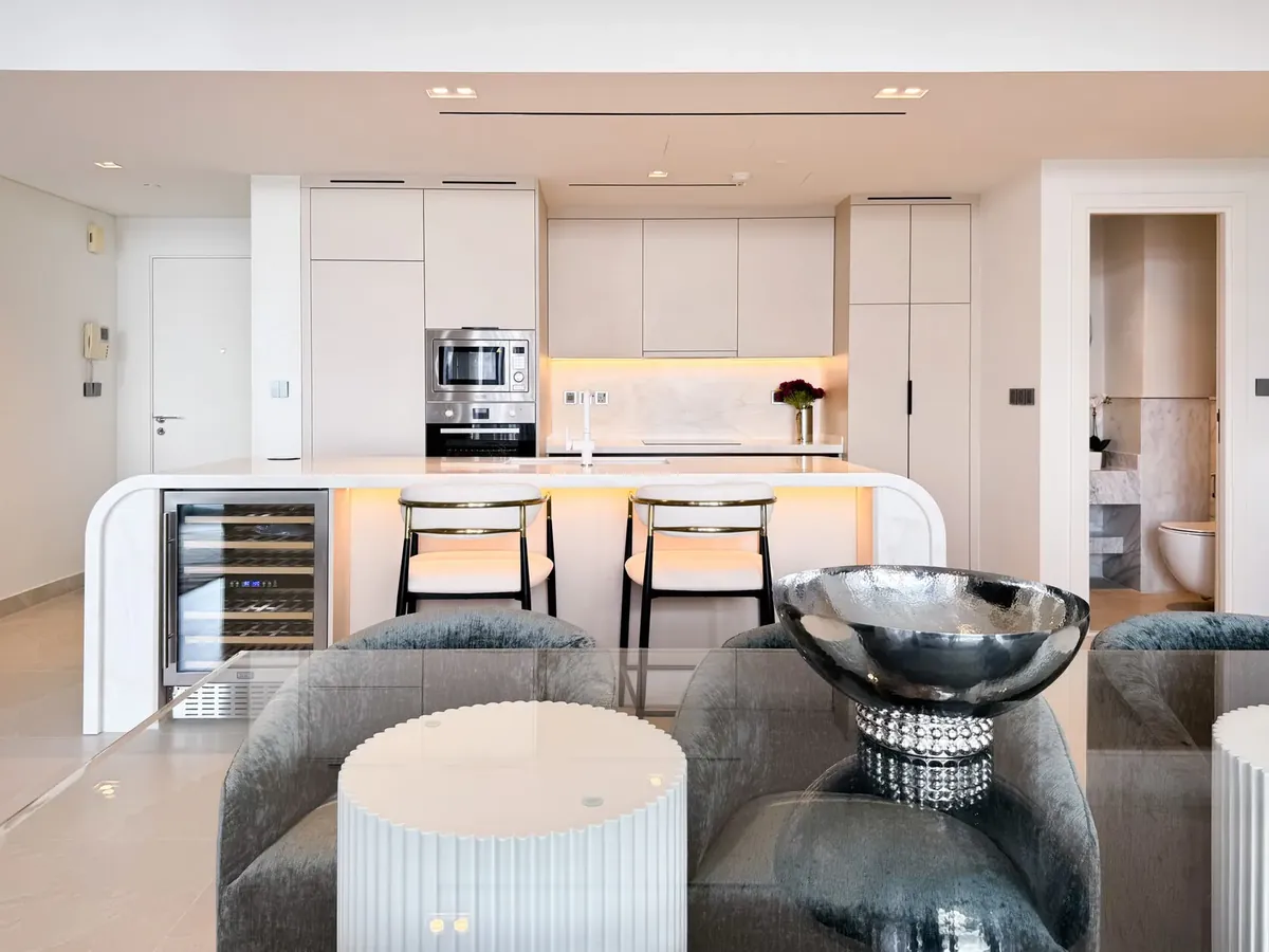 Open-plan Dubai apartment with white kitchen, illuminated curved island with bar stools and grey sofas in the foreground